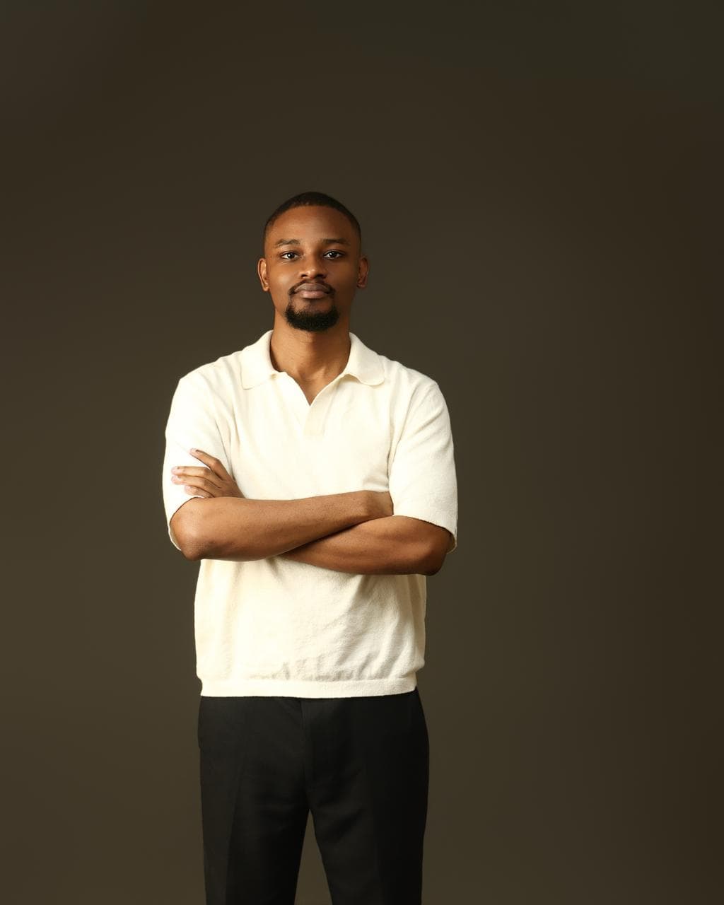 Kehinde Abiuwa standing with his arms folded, wearing a cream knit polo